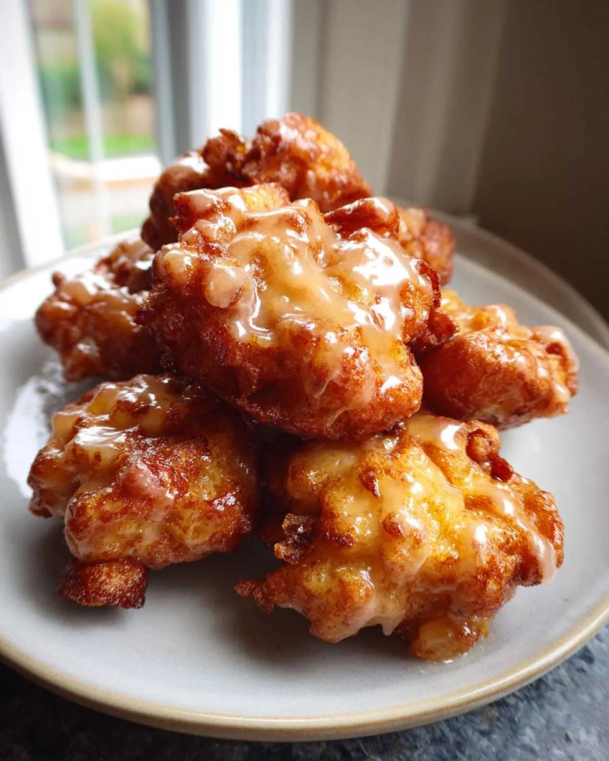 A stack of golden brown, glazed apple fritter bites piled high on a light-colored plate.