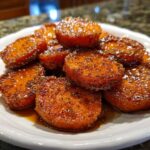Close-up of shiny, glazed candied sweet potatoes slices piled high on a white plate, coated in spices.