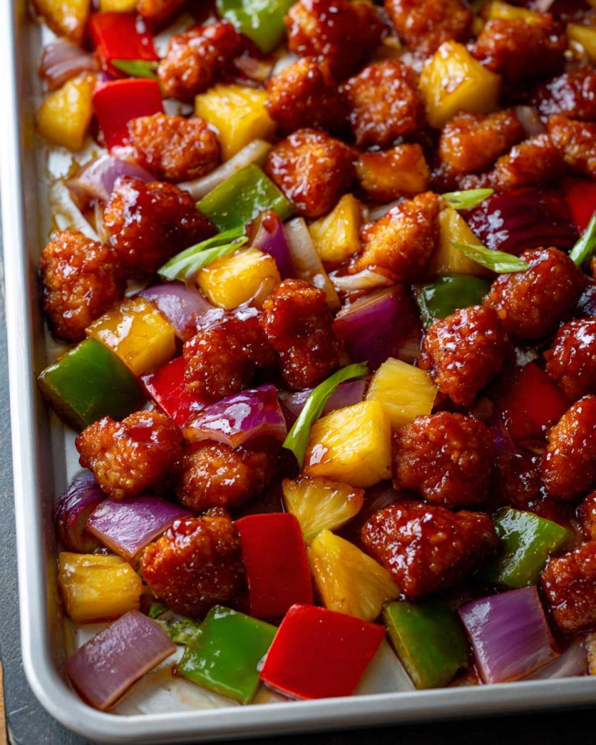 Close-up of glazed Hawaiian chicken pieces mixed with pineapple, red onion, and bell peppers on a sheet pan.