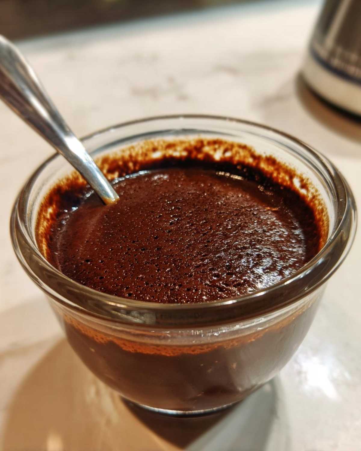 Close-up of rich, dark homemade chocolate pudding with a slightly foamy top in a small glass bowl with a spoon.