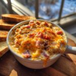 A close-up of a hot, cheesy reuben dip topped with spices, served in a white ramekin next to toasted bread.