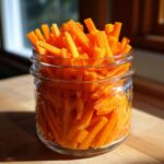 A clear glass jar filled with bright orange, julienned pickled carrots sticks, sitting on a wooden surface.