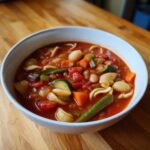 A close-up of a white bowl filled with vibrant minestrone soup, featuring chunks of tomato, zucchini, carrots, green beans, and small pasta shells.