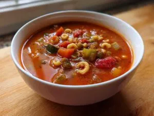 Close-up of a white bowl filled with rich, red minestrone soup featuring diced vegetables and small pasta shapes.