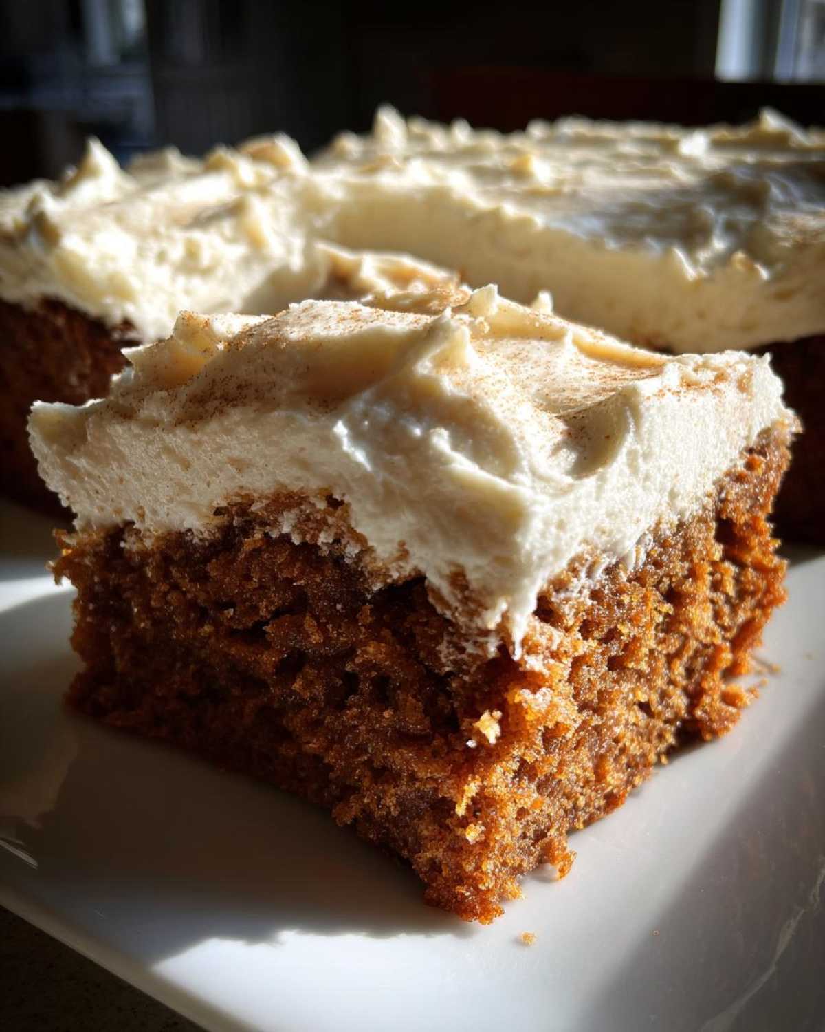A close-up slice of moist, dark applesauce cake topped with thick cream cheese frosting dusted with cinnamon.