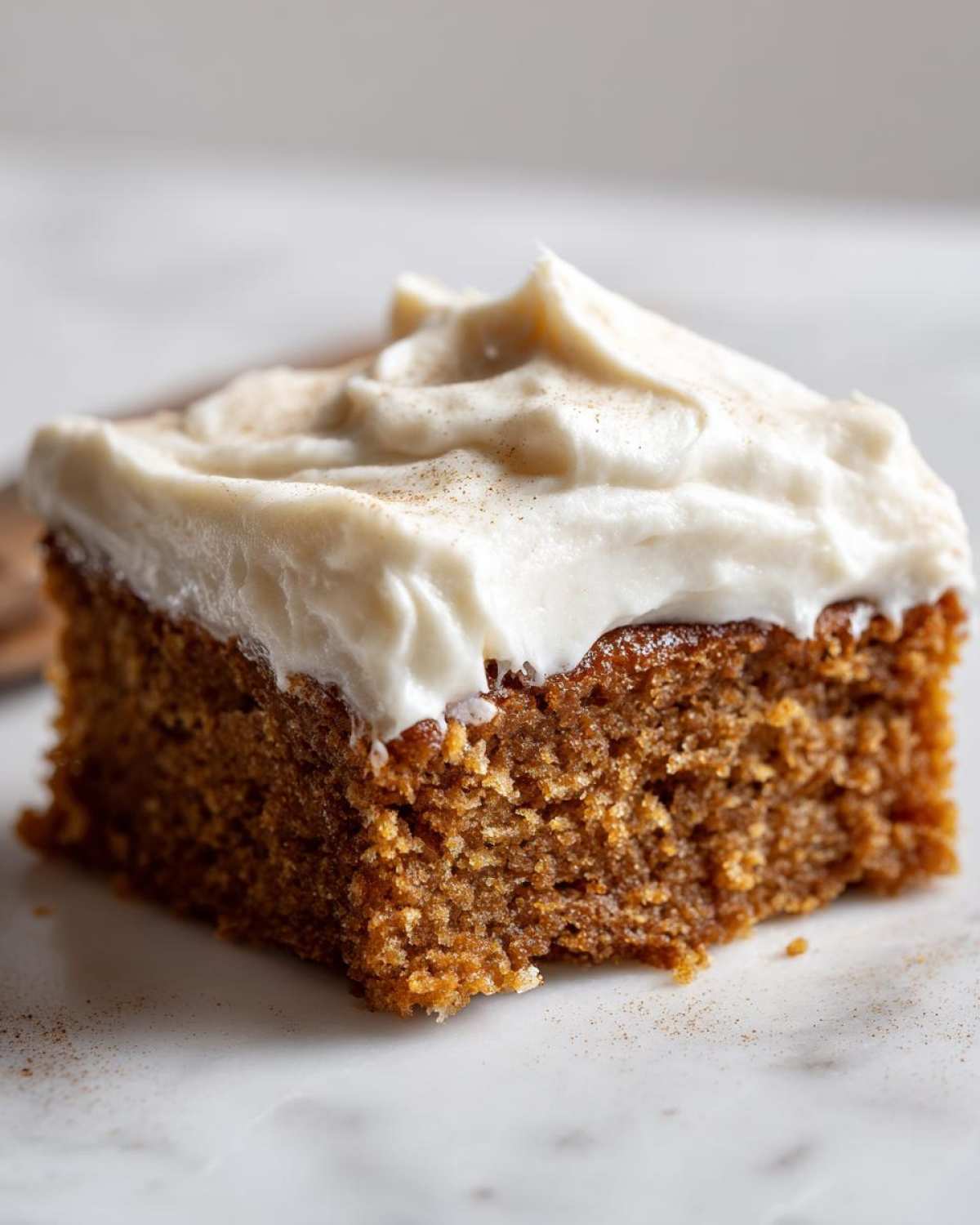 Close-up of a moist square slice of applesauce cake topped with thick white frosting and a dusting of spice.