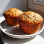 Two golden brown, moist bran muffins sitting on a light ceramic plate, illuminated by sunlight.