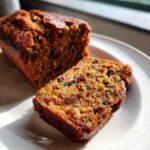 Close-up of a moist slice of fruit cake, rich with raisins and candied peel, next to the main loaf on a white plate.