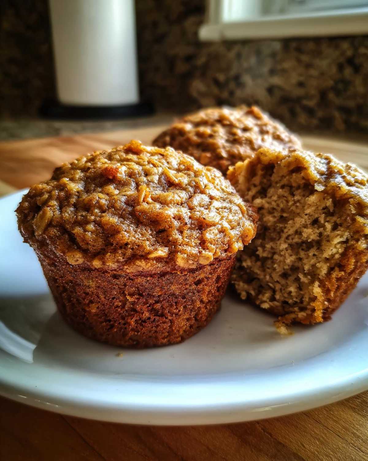 Three moist oatmeal muffins on a white plate, one broken open to show the texture.