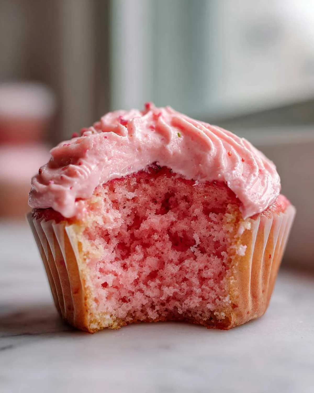Close-up of a strawberry cupcake with a bite taken out, showing the moist pink crumb and pink strawberry frosting.