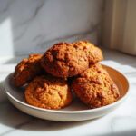 A close-up of a small stack of freshly baked, golden brown persimmon cookies resting on a light ceramic plate.