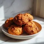 A close-up of a small stack of freshly baked, golden brown persimmon cookies resting on a light ceramic plate.