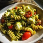 A vibrant bowl of pesto pasta salad featuring fusilli pasta, cherry tomatoes, and small mozzarella balls coated in green pesto.