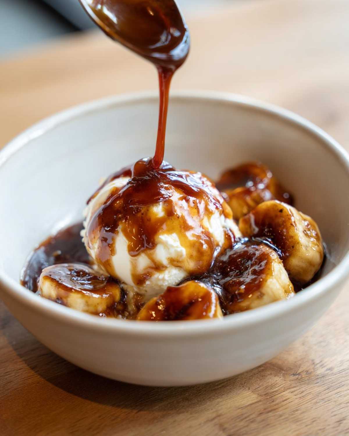 A spoonful of rich caramel sauce is poured over vanilla ice cream and caramelized bananas in a bowl of Bananas Foster.