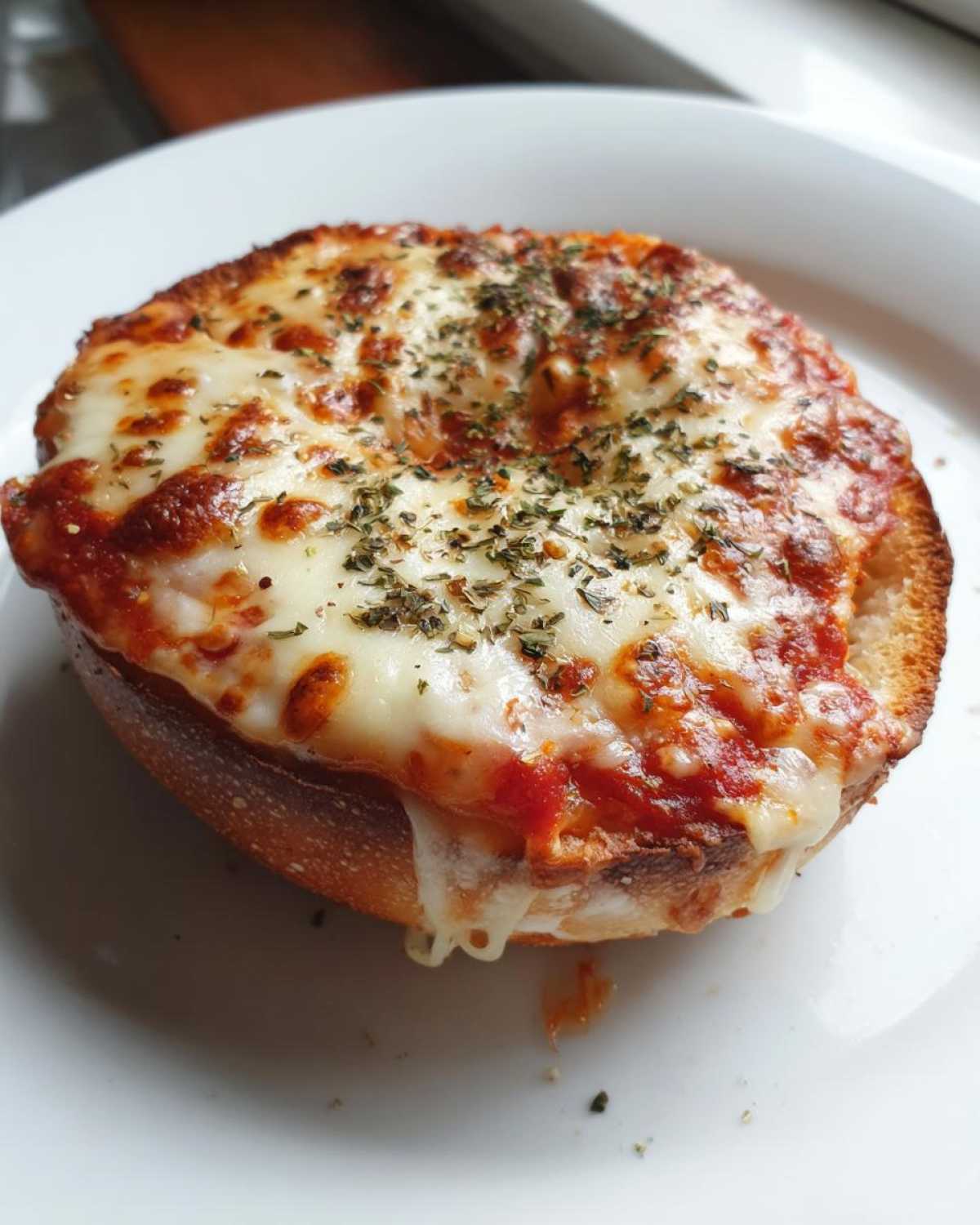 Close-up of a freshly baked pizza bagel half topped with melted mozzarella, tomato sauce, and dried herbs.