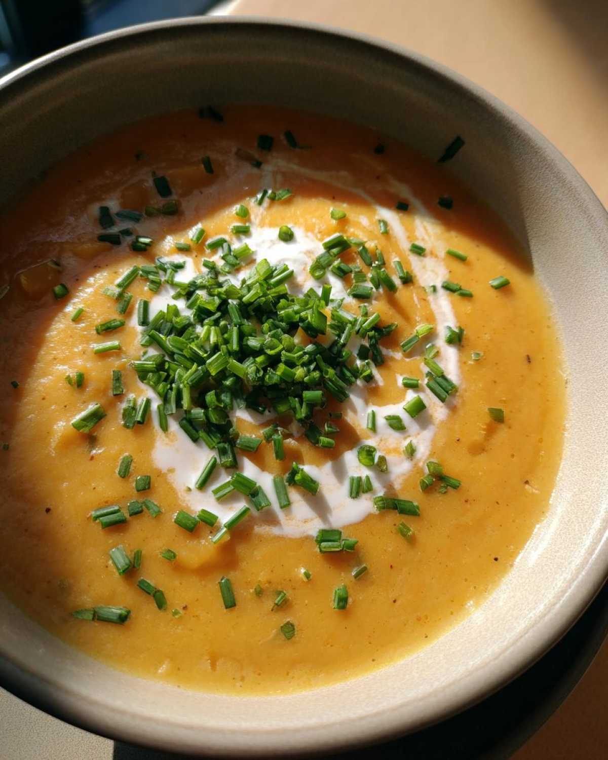 Close-up of a vibrant orange cheese soup topped with a swirl of cream and fresh chopped chives.