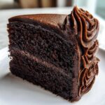 A close-up of a moist slice of dark chocolate cake recipe topped with smooth and piped chocolate frosting.