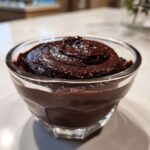 A close-up of rich, dark homemade chocolate pudding swirled in a small, clear glass serving dish.