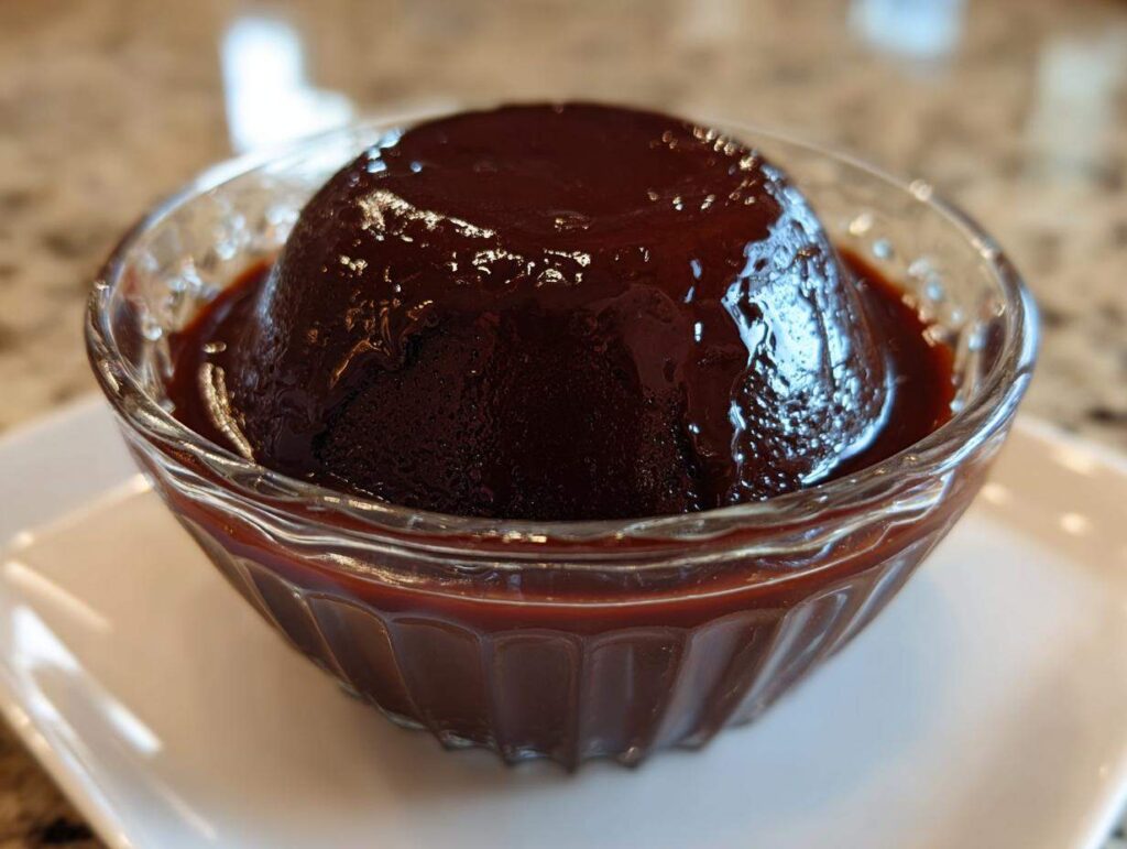 A rich, dark mound of homemade chocolate pudding served in a fluted glass bowl with extra sauce.