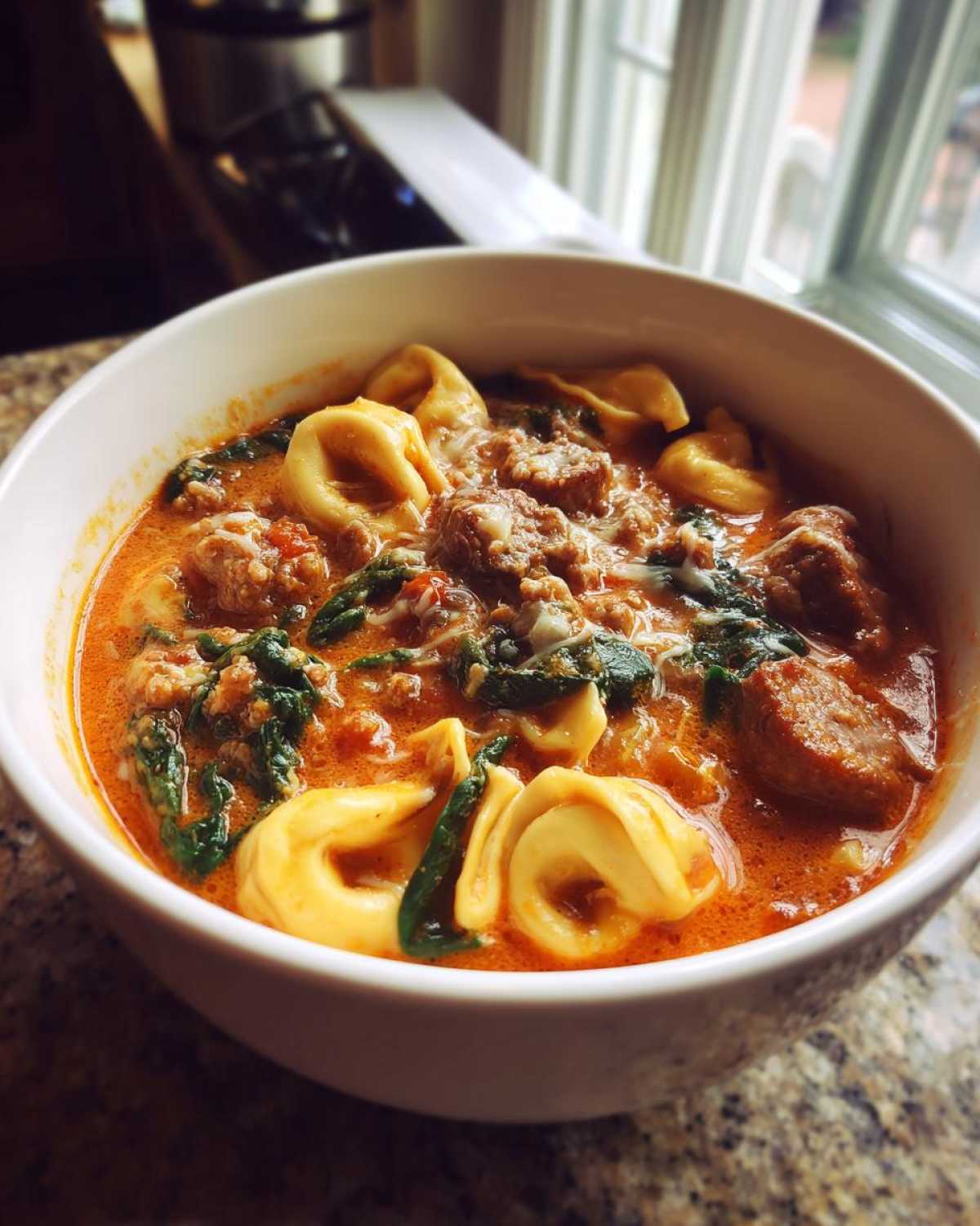Close-up of a white bowl filled with creamy sausage tortellini soup, featuring chunks of sausage, spinach, and cheese.