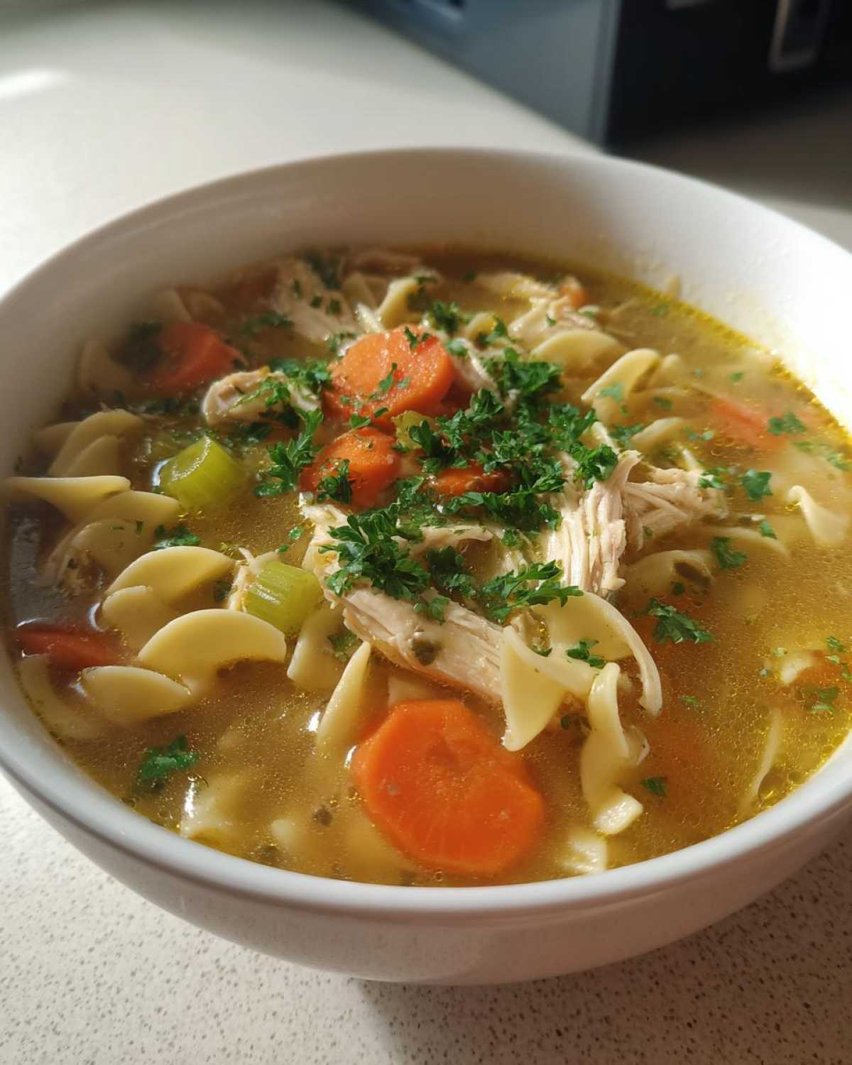 Close-up of a white bowl filled with steaming slow cooker chicken noodle soup, featuring egg noodles, shredded chicken, carrots, and parsley.