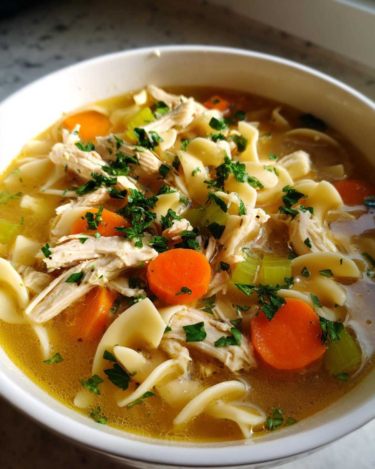 A close-up, appetizing bowl of slow cooker chicken noodle soup showing shredded chicken, egg noodles, carrots, celery, and fresh parsley.