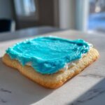 Close-up of a square sugar cookie topped with white frosting and vibrant blue sugar cookie icing recipe.