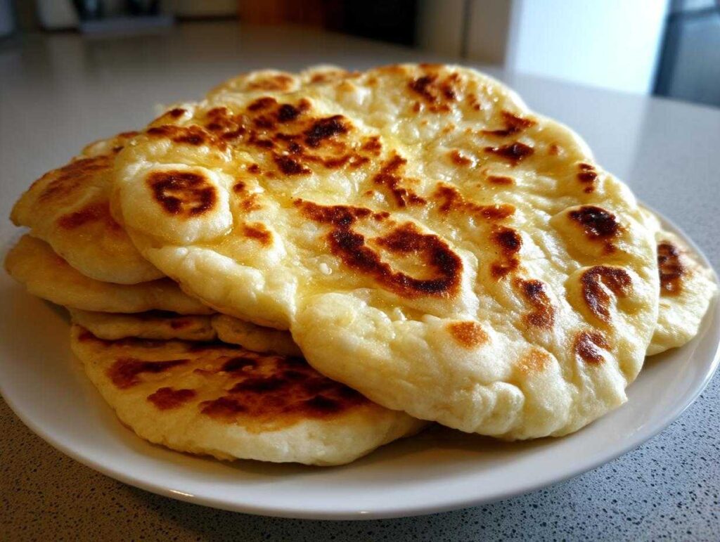 A stack of freshly made, fluffy naan bread rounds with golden-brown charred spots, glistening with butter.