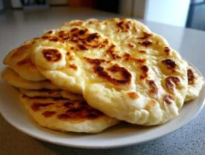 A stack of freshly made, fluffy naan bread rounds with golden-brown charred spots, glistening with butter.