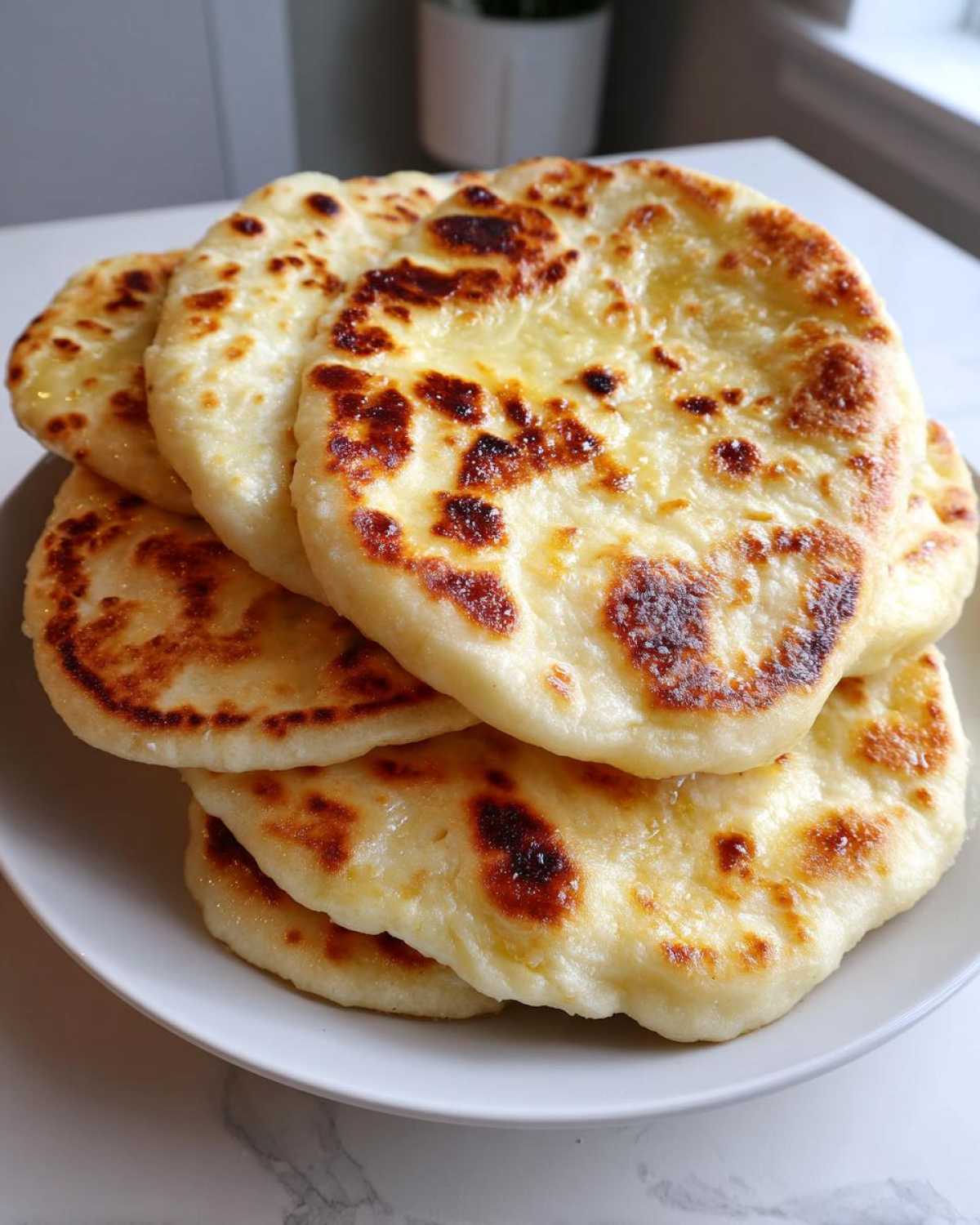 A stack of freshly cooked, golden-brown naan bread rounds piled on a white plate.