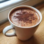 A white mug filled with rich, frothy hot chocolate dusted with cocoa powder, sitting on a wooden surface.
