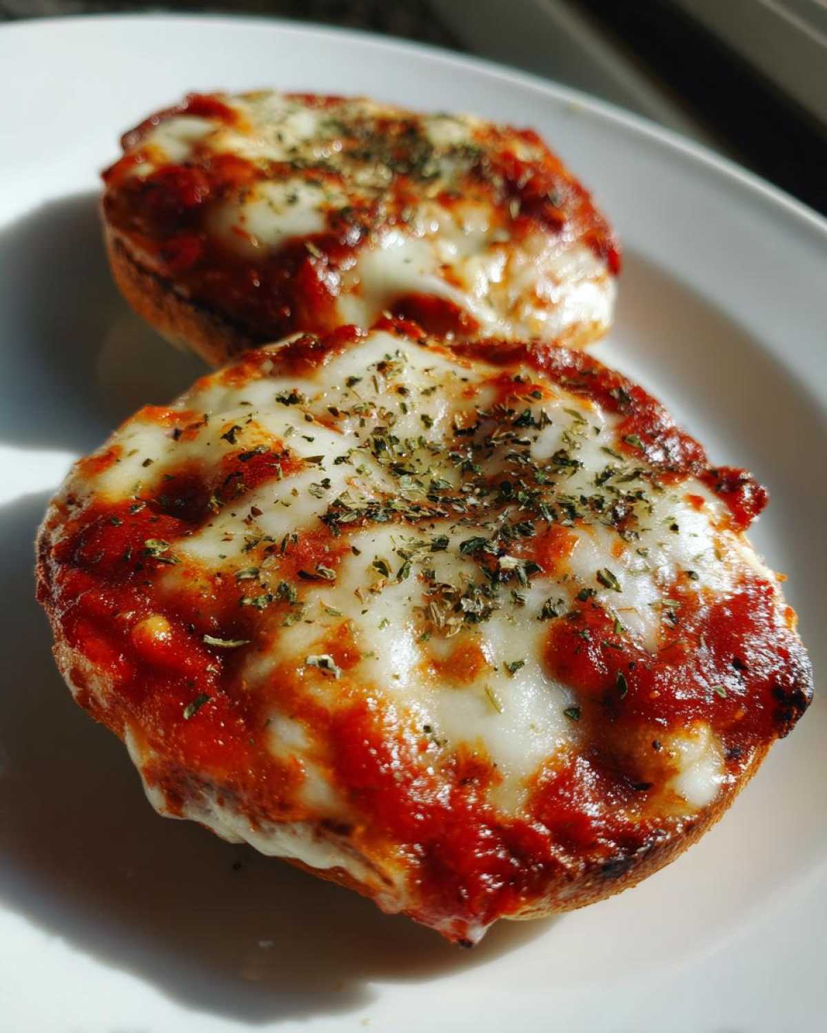Close-up of two hot pizza bagels topped with melted mozzarella, rich tomato sauce, and dried herbs.