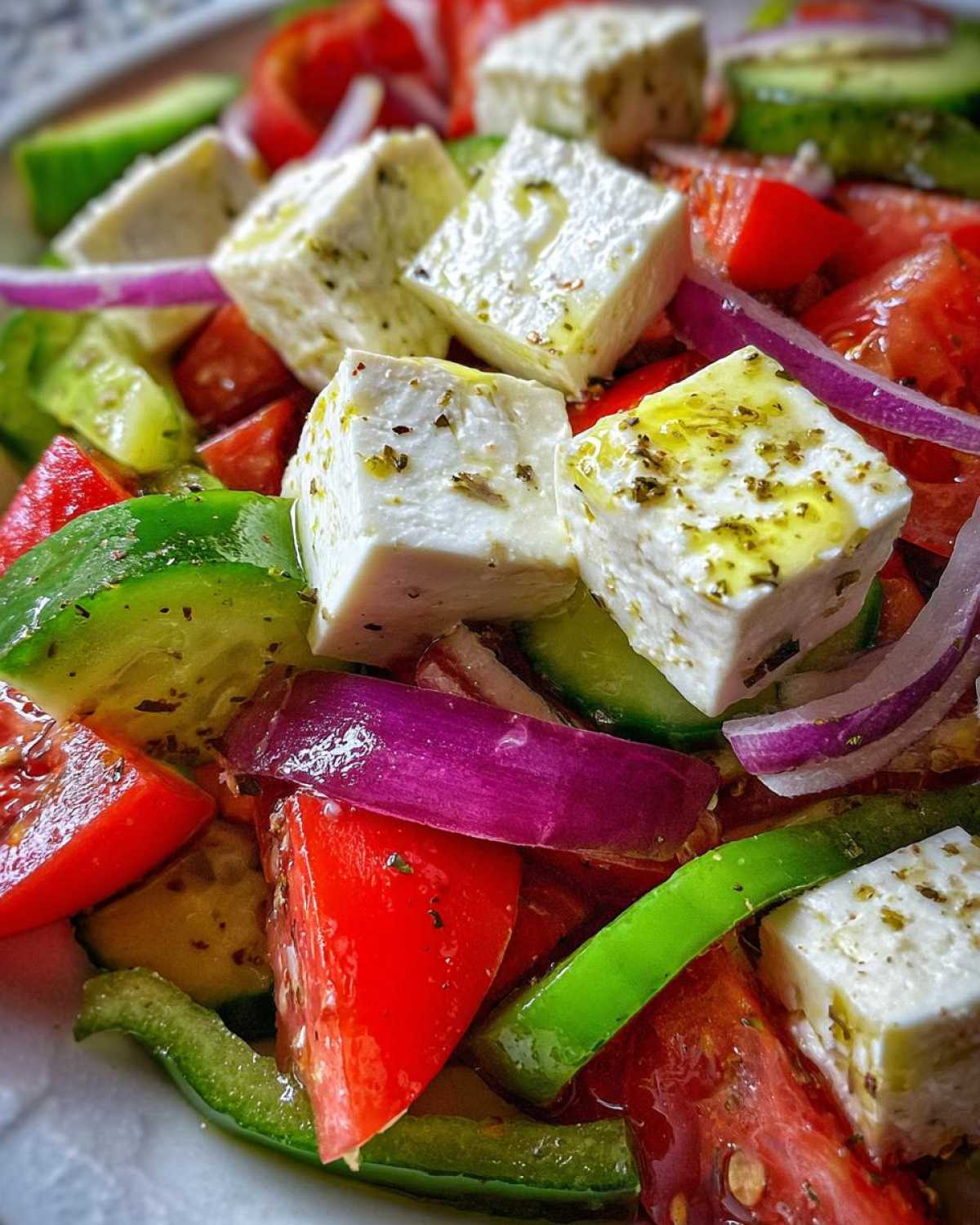 Close-up of a vibrant greek salad featuring large cubes of feta cheese drizzled with olive oil and herbs, mixed with tomatoes, cucumbers, and red onion.