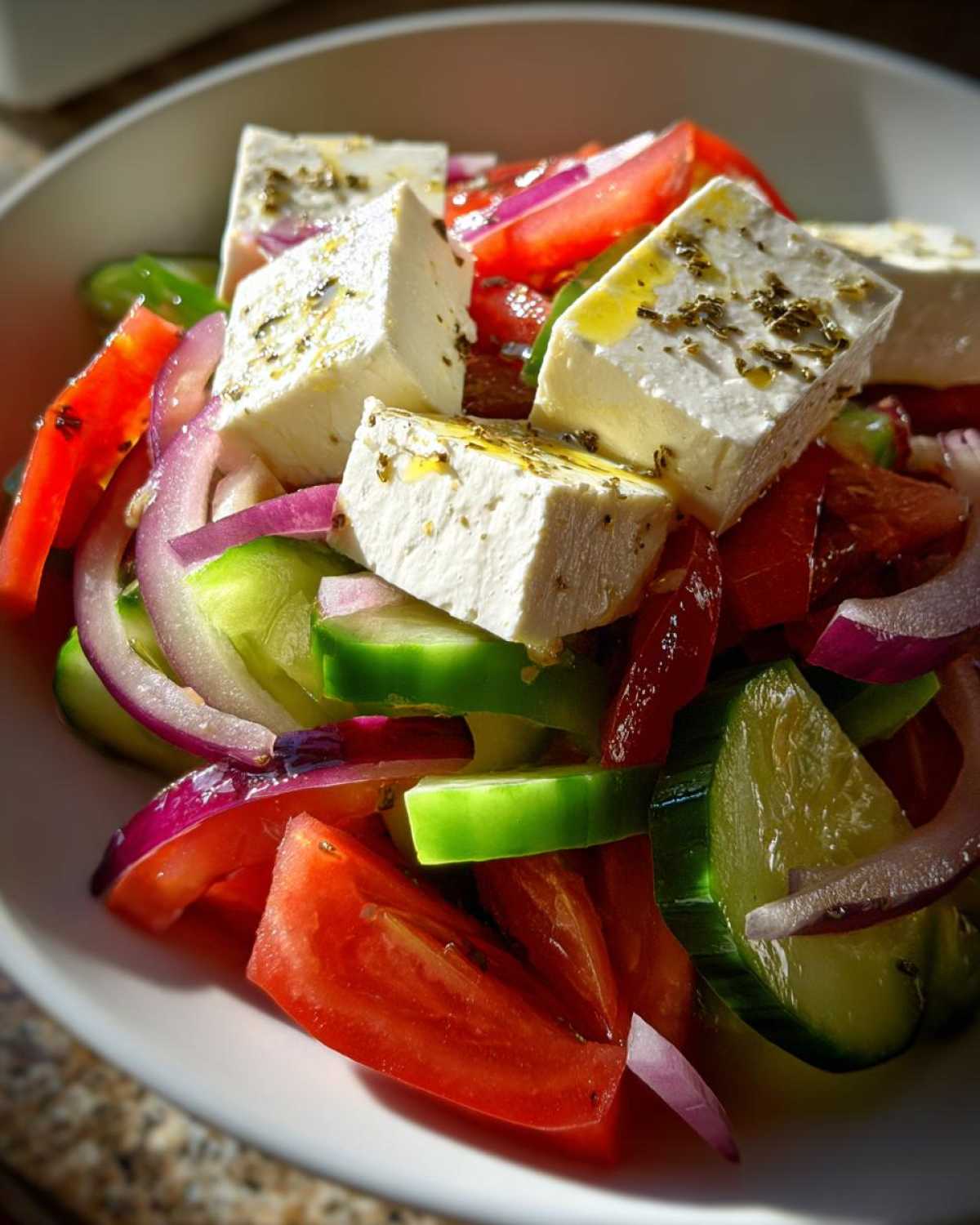 Close-up of a vibrant greek salad featuring large cubes of feta cheese drizzled with oil and herbs, mixed with tomatoes, cucumbers, and red onion.