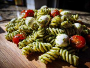 Close-up of vibrant pesto pasta salad featuring fusilli, cherry tomatoes, and small mozzarella balls on a wooden board.