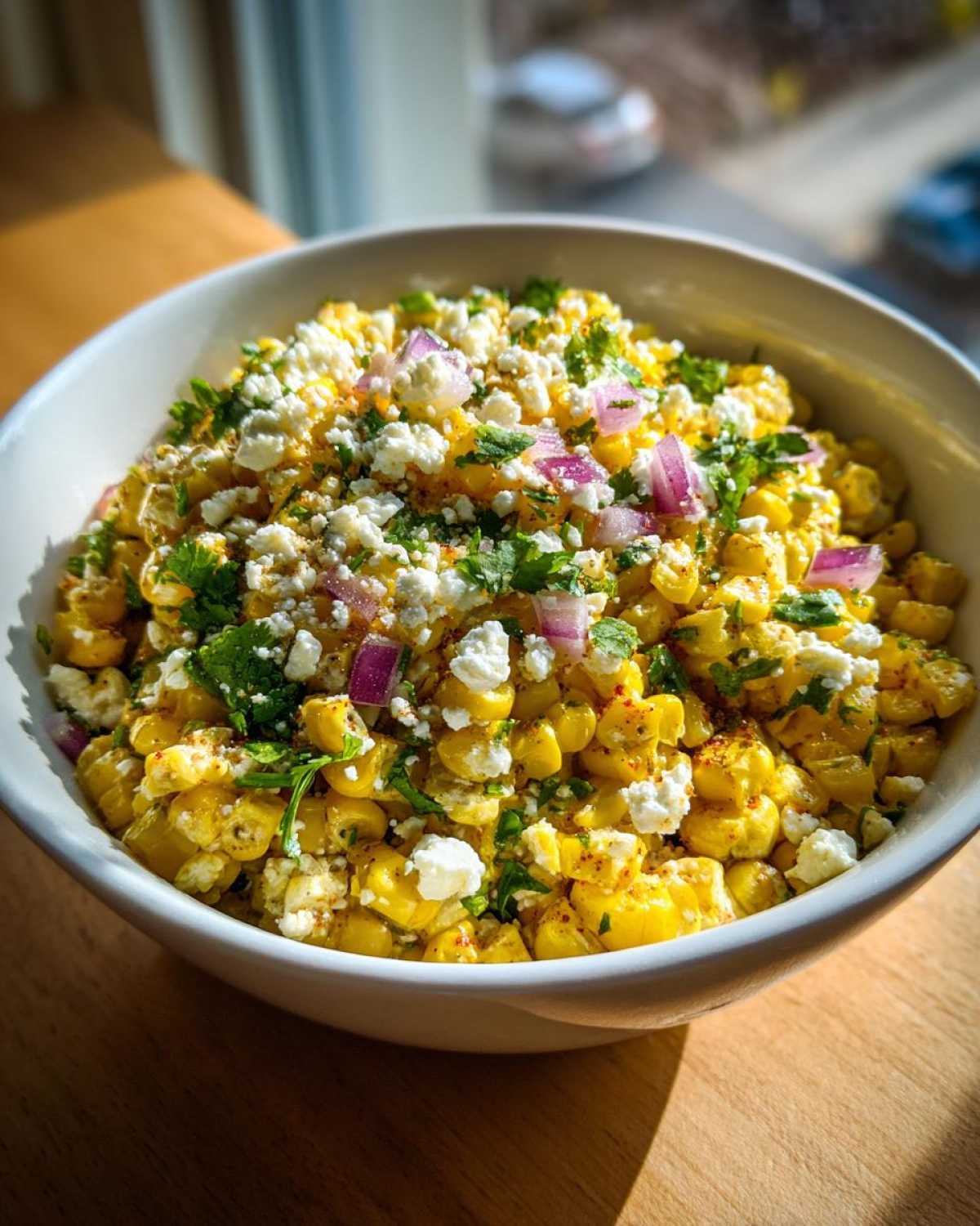 A close-up of a white bowl filled with bright yellow street corn salad topped with cotija cheese, red onion, and cilantro.