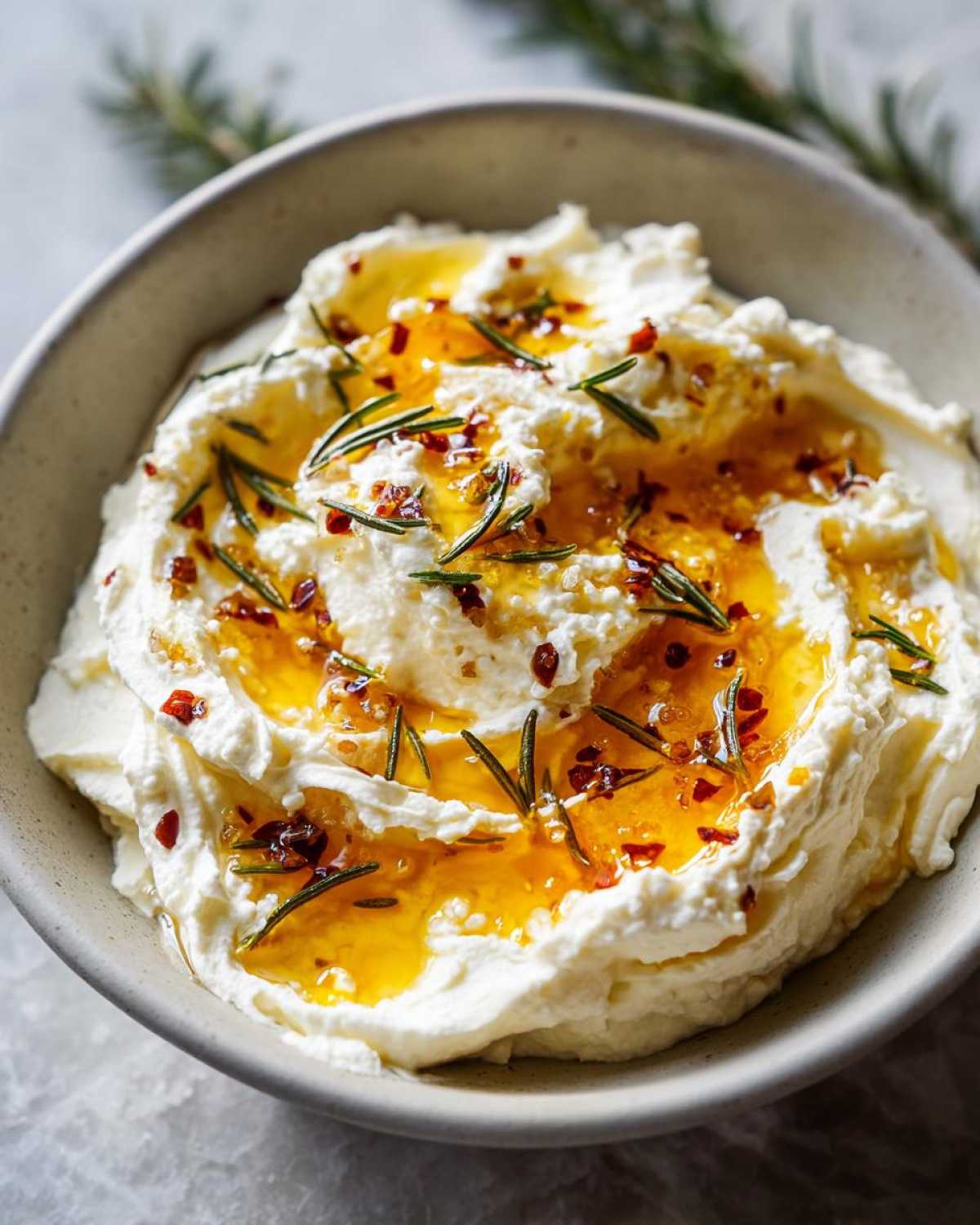 Close-up of creamy whipped ricotta dip drizzled with honey, chili flakes, and fresh rosemary sprigs.