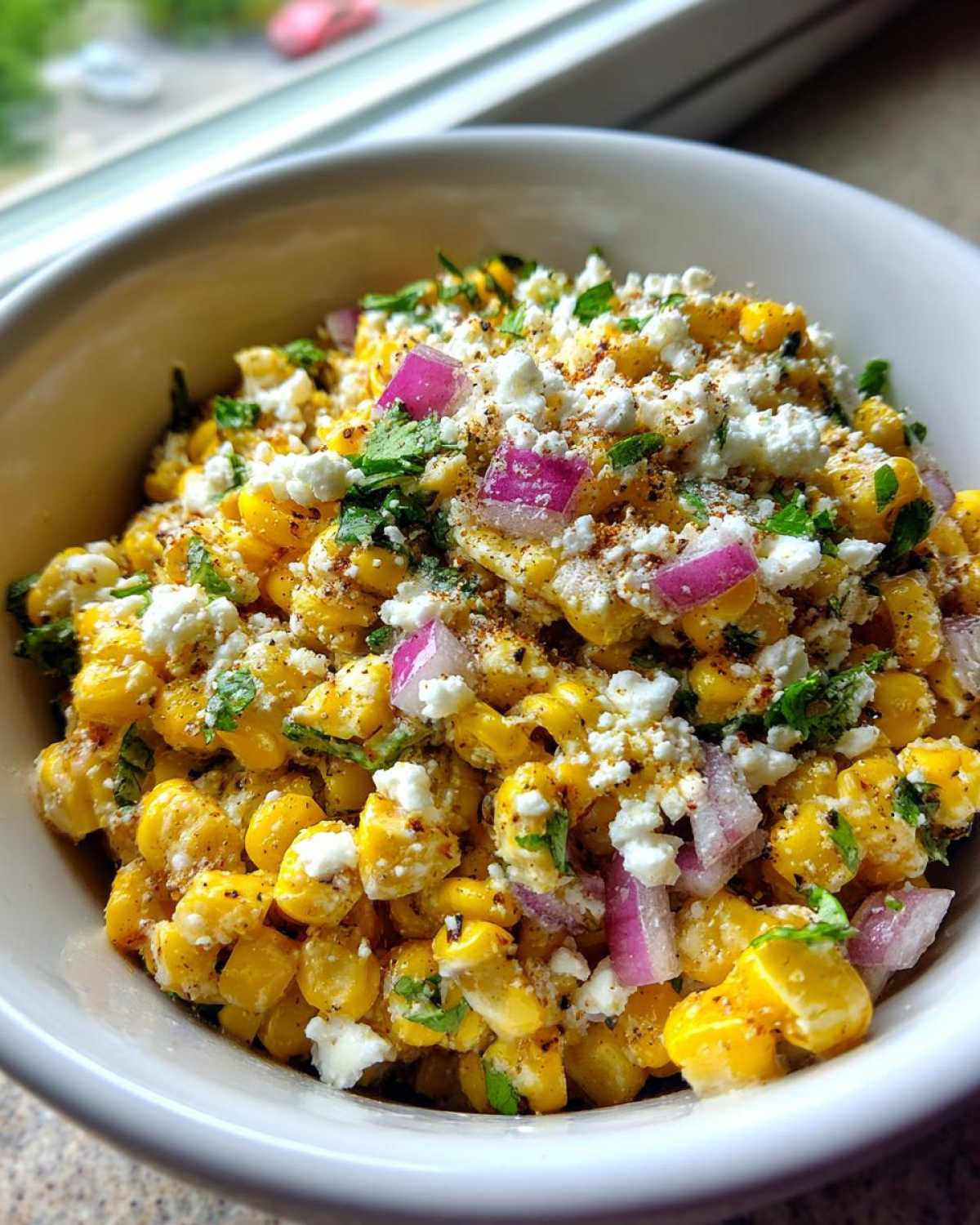 Close-up of a white bowl filled with vibrant street corn salad featuring corn kernels, crumbled white cheese, red onion, and cilantro.