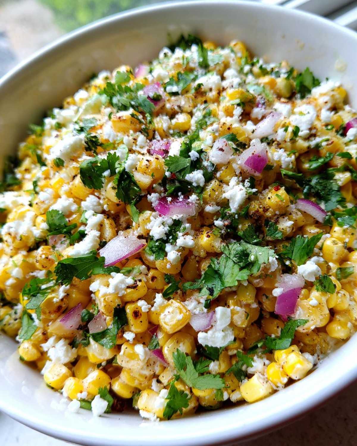Close-up of a white bowl filled with vibrant street corn salad, topped with cotija cheese, red onion, and cilantro.