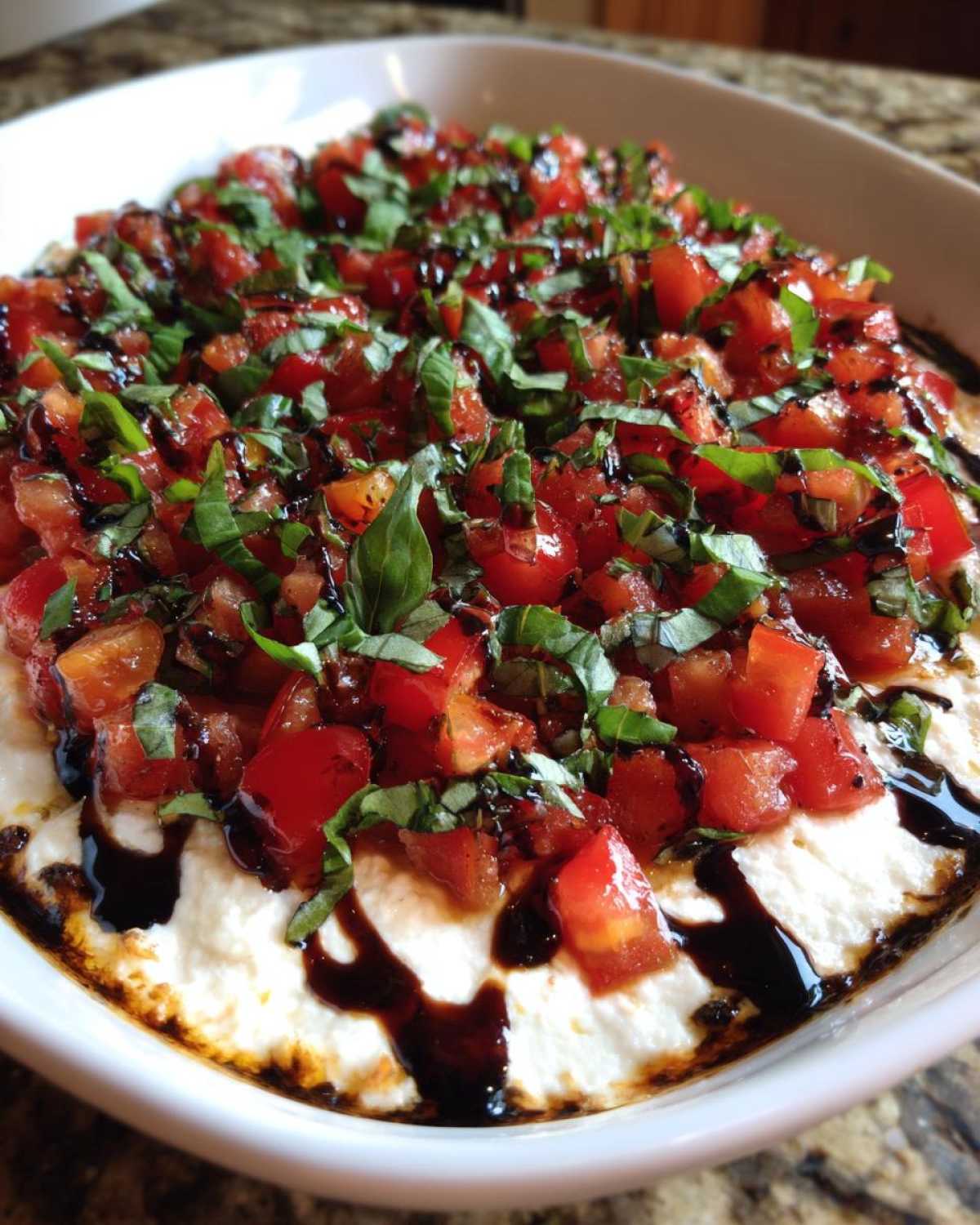 Close-up of a creamy bruschetta dip topped with diced tomatoes, fresh basil, and a balsamic glaze drizzle.
