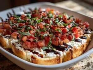 A close-up of a warm bruschetta dip served in a white dish, featuring layers of toasted bread, creamy cheese, diced tomatoes, and fresh basil.