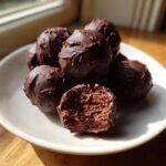 A pile of rich, chocolate-coated oreo balls on a white plate, one is bitten open showing the dark filling.