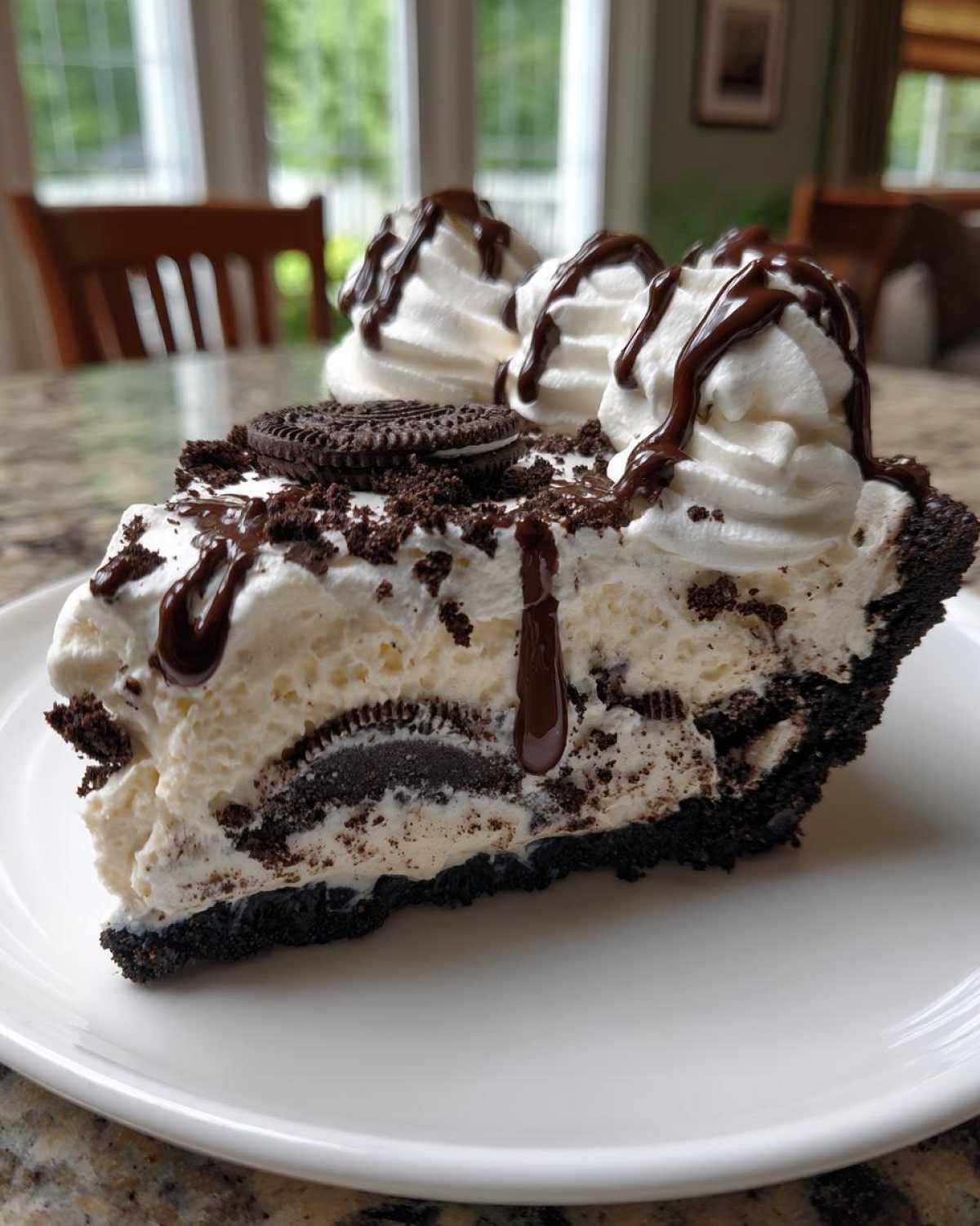 A decadent slice of ice cream pie featuring a dark cookie crust, creamy filling with cookie chunks, topped with whipped cream and chocolate syrup.
