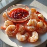 A white plate featuring perfectly cooked shrimp cocktail pieces arranged around a small glass bowl of zesty red cocktail sauce.