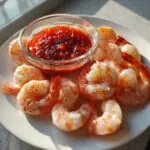 A white plate featuring perfectly cooked shrimp cocktail pieces arranged around a small glass bowl of zesty red cocktail sauce.
