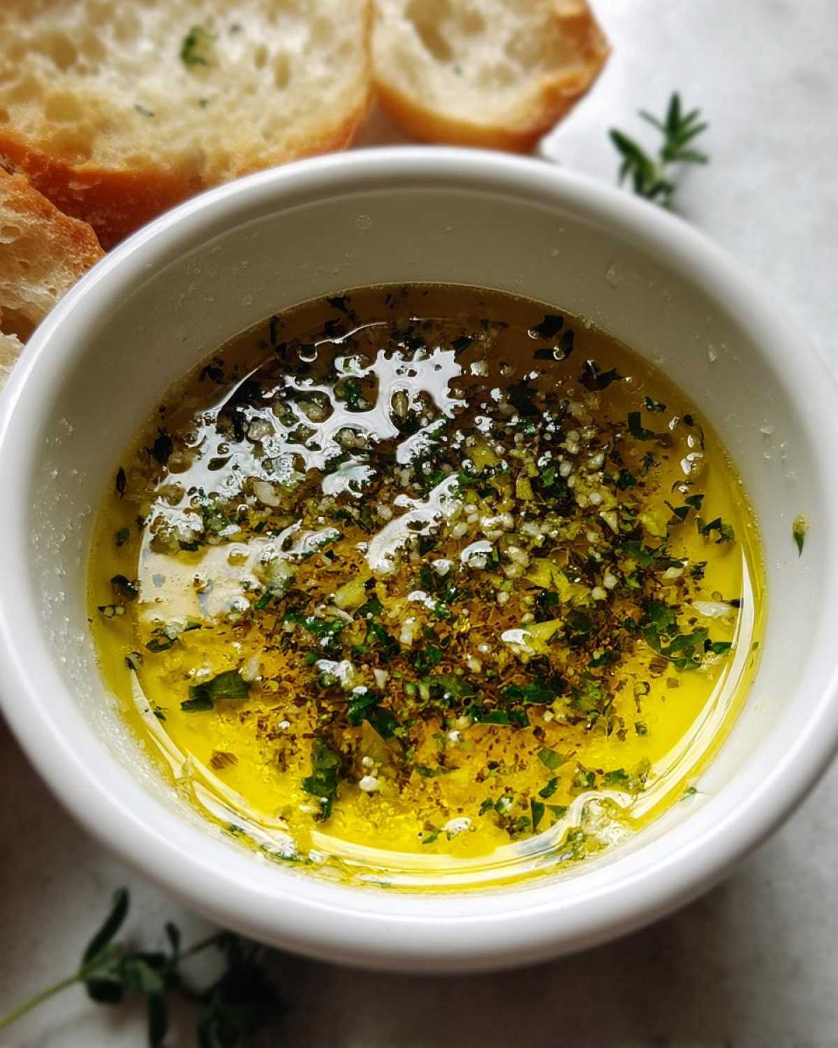 Close-up of a white bowl filled with bright yellow olive oil bread dip seasoned with garlic and herbs.
