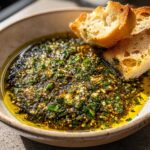 Close-up of a rustic bowl filled with vibrant green olive oil bread dip, topped with herbs and spices, served with toasted bread pieces.