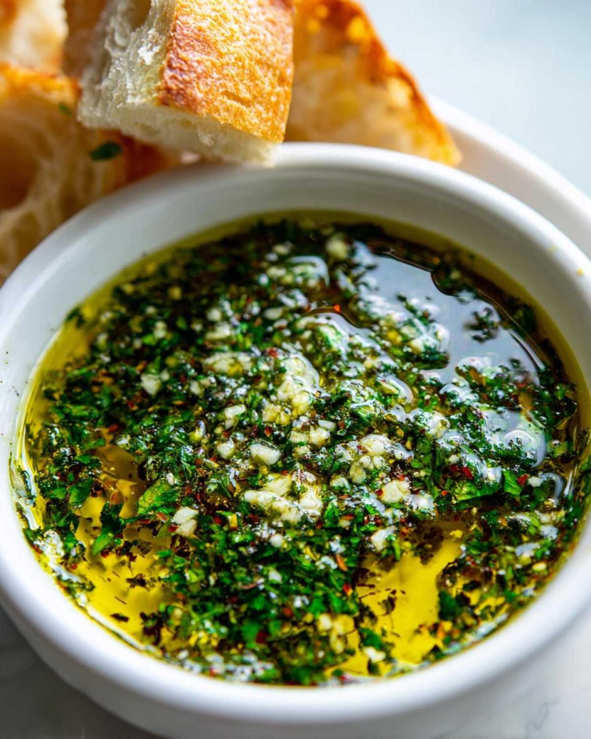 Close-up of a white bowl filled with vibrant olive oil bread dip featuring herbs and garlic, with crusty bread pieces above.