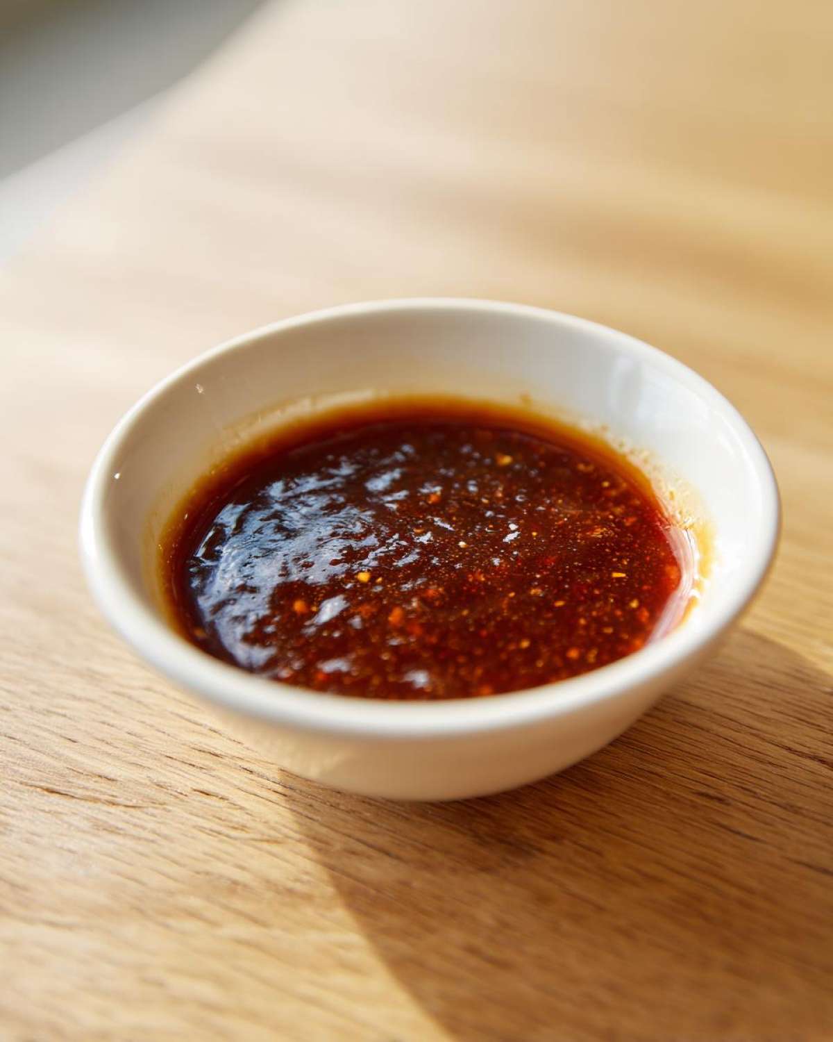 A close-up of thick, dark red homemade bbq sauce speckled with spices, served in a small white dipping bowl on a wooden surface.