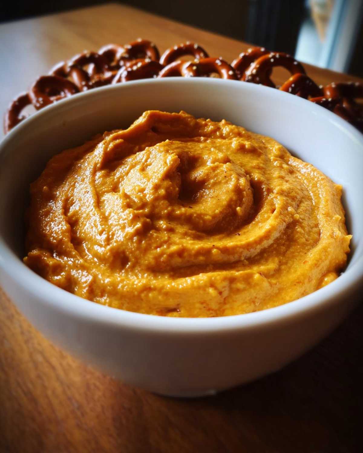 Close-up of a bowl of vibrant orange beer cheese dip, served with soft pretzels blurred in the background.